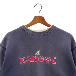May include: A navy blue KANGOL sweatshirt, featuring the brand name in red with a white outline, and a kangaroo logo. The text "CUMBRIA, ENGLAND SINCE 1938" is below the logo. The sweatshirt is displayed on a wooden hanger.
