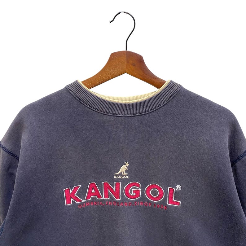 May include: A navy blue KANGOL sweatshirt, featuring the brand name in red with a white outline, and a kangaroo logo. The text "CUMBRIA, ENGLAND SINCE 1938" is below the logo. The sweatshirt is displayed on a wooden hanger.
