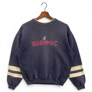 May include: A navy blue Kangol sweatshirt with the brand name in red and a kangaroo logo. The sweatshirt has cream-coloured stripes on the sleeves and a cream-coloured collar. The sweatshirt is hanging on a wooden hanger.