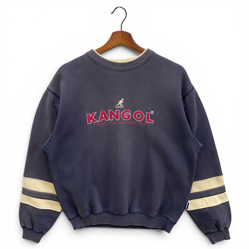 May include: A navy blue Kangol sweatshirt with the brand name in red and a kangaroo logo. The sweatshirt has cream-coloured stripes on the sleeves and a cream-coloured collar. The sweatshirt is hanging on a wooden hanger.