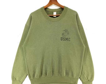 May include: Olive green sweatshirt with the text "USMC" and a Marine Corps emblem. The crew neck sweatshirt is hanging on a wooden hanger. The garment is a solid color and has ribbed cuffs and hem.