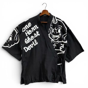 May include: Black kimono-style robe with white devil illustrations and the text "One Horn Ghost Devil." The robe is suspended on a wooden hanger. The robe features a wrap-around design.