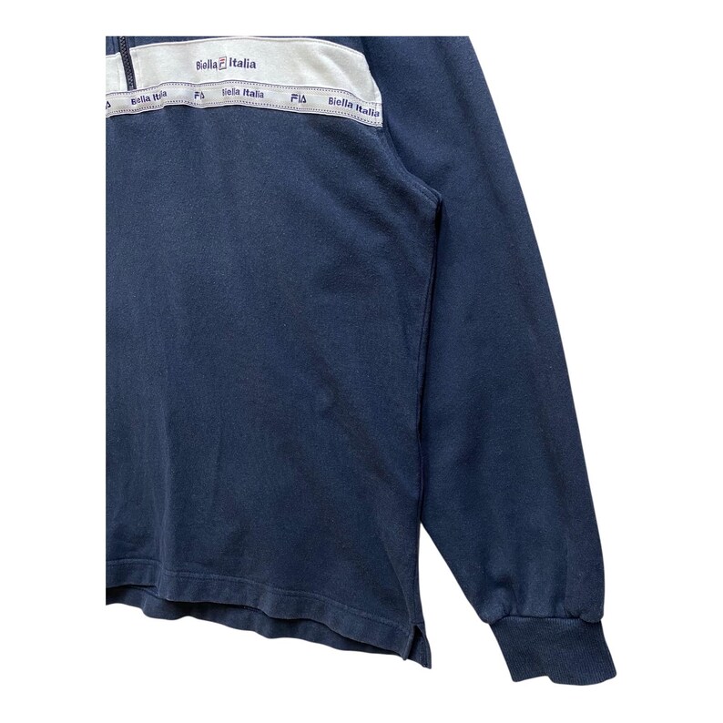 May include: A navy blue long-sleeved shirt with a white and grey panel across the chest. The panel has the text "Bella Italia" repeated. The shirt has a ribbed cuff and a small split at the hem.