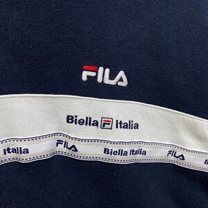 May include: Navy blue sweatshirt with a white band across the chest. The band features the text "Biella Italia" and "FILA" in navy blue. The FILA logo is embroidered in white with red accents.