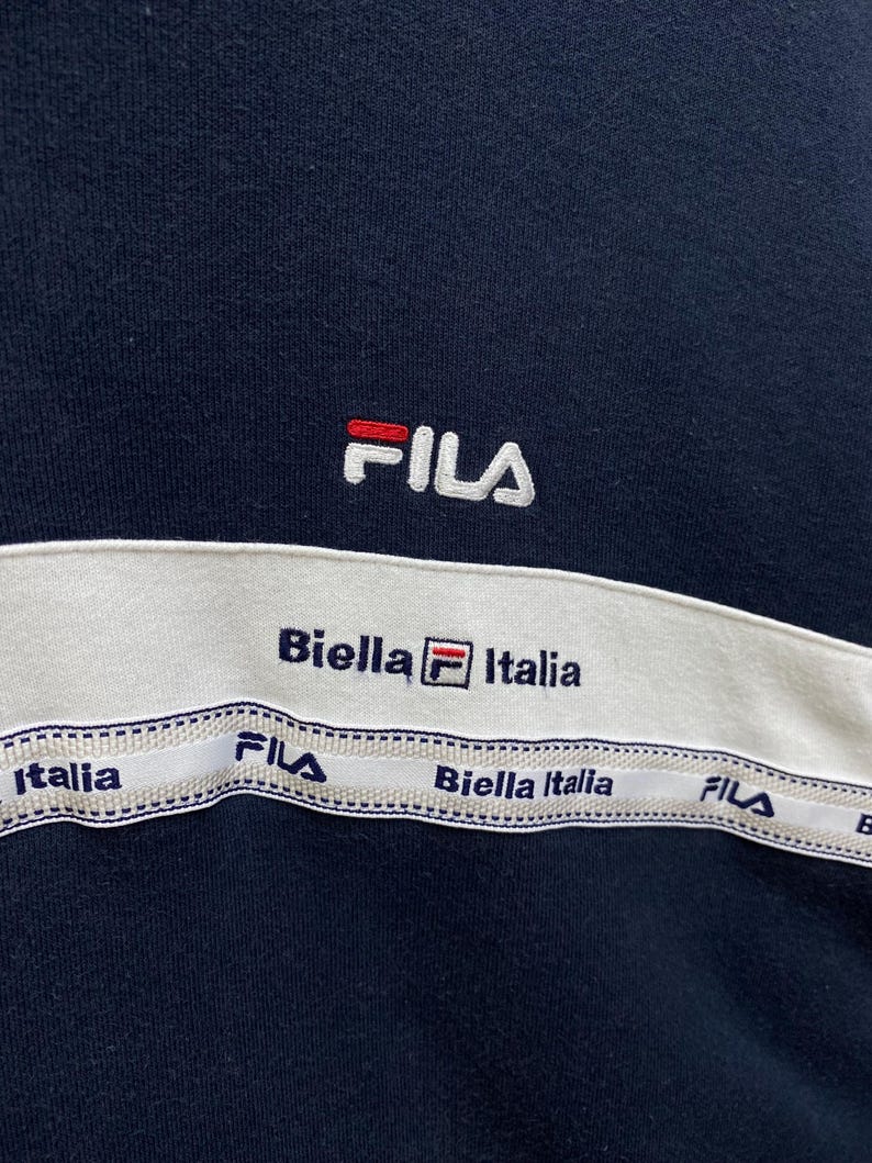 May include: Navy blue sweatshirt with a white band across the chest. The band features the text "Biella Italia" and "FILA" in navy blue. The FILA logo is embroidered in white with red accents.