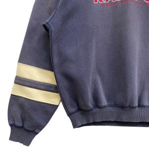 May include: A navy blue sweatshirt with cream stripes on the left sleeve. The sweatshirt features a ribbed hem and cuffs. The text "CUMBRIA ENGLAND SINCE" is visible in red on the upper right side.