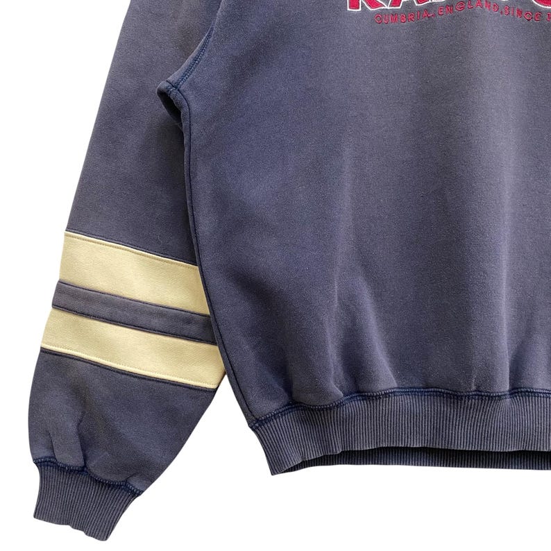 May include: A navy blue sweatshirt with cream stripes on the left sleeve. The sweatshirt features a ribbed hem and cuffs. The text "CUMBRIA ENGLAND SINCE" is visible in red on the upper right side.