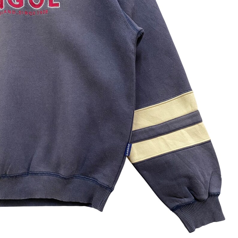 May include: A navy blue sweatshirt with the word "KANGOL" in red lettering. The sleeve features two beige stripes. The sweatshirt has a ribbed hem and cuffs. The text "BRAND SINCE 1938" is above the word "KANGOL".