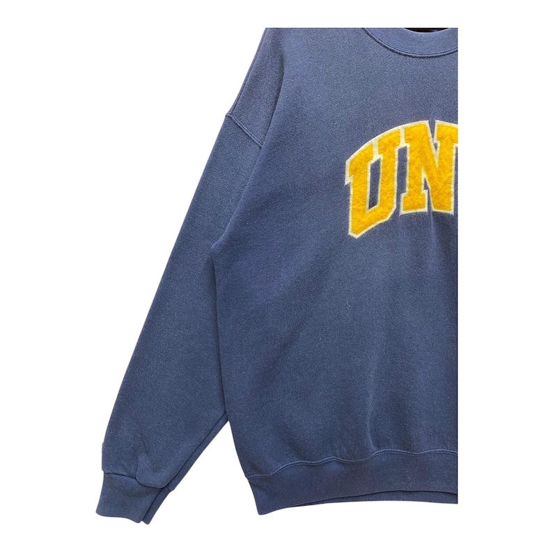 Op de afbeelding: Een blauwe sweatshirt met het woord "UN" in geel, omlijnd met wit. De sweatshirt heeft een ronde hals en lange mouwen. De stof lijkt een zacht, comfortabel materiaal te zijn.