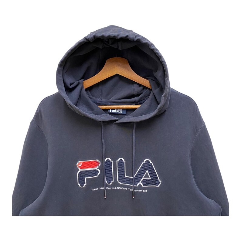 May include: A navy blue hooded sweatshirt with the brand name "FILA" in a pixelated design across the chest. The logo features red and white accents. The sweatshirt has a drawstring and a wooden hanger.