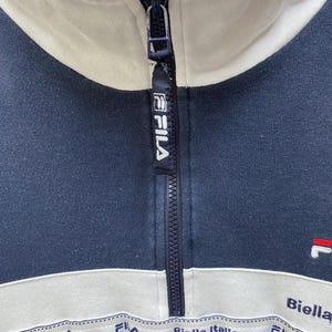 May include: Navy blue athletic jacket with a white collar and stripe across the chest. The jacket features a zip closure and the brand name "FILA" on the zip pull. The words "Biella Italia" are printed on the white stripe.