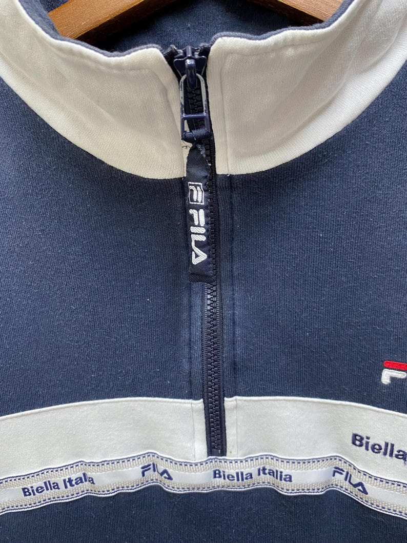 May include: Navy blue athletic jacket with a white collar and stripe across the chest. The jacket features a zip closure and the brand name "FILA" on the zip pull. The words "Biella Italia" are printed on the white stripe.