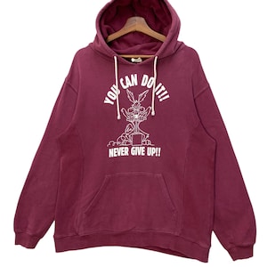 May include: A maroon hoodie with a kangaroo pocket and a drawstring hood. The front displays a white graphic of a cartoon coyote with the text "YOU CAN DO IT!!" above and "NEVER GIVE UP!!" below.
