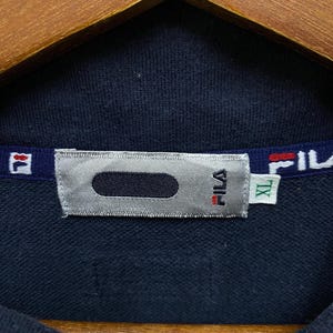May include: A close-up of a navy blue FILA zip-up jacket. The collar displays the FILA logo in red and white. A silver tag with the FILA logo and an XL size tag are visible. The jacket is hanging on a wooden hanger.