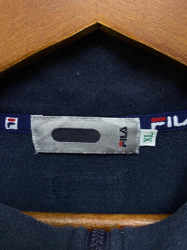 May include: A close-up of a navy blue FILA zip-up jacket. The collar displays the FILA logo in red and white. A silver tag with the FILA logo and an XL size tag are visible. The jacket is hanging on a wooden hanger.