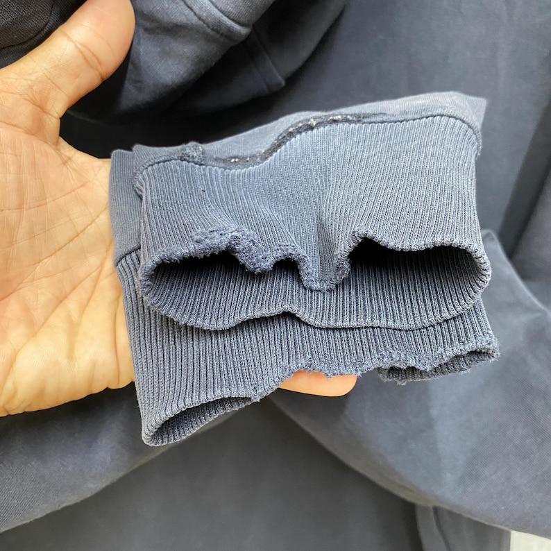 May include: Close-up of a dark blue hoodie sleeve cuff. The cuff is ribbed and has a distressed, frayed edge. The fabric appears to be a soft, worn material. The image focuses on the texture and detail of the garment.