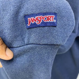 Op de afbeelding: Close-up van een blauwe sweatshirtmouw met een geribbelde manchet. Een rechthoekige patch met de merknaam "JANSPORT" in rode letters op een witte achtergrond is op de mouw genaaid.