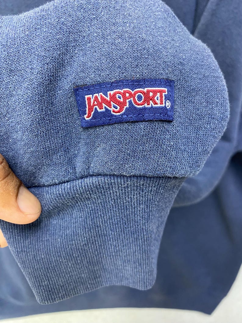 Op de afbeelding: Close-up van een blauwe sweatshirtmouw met een geribbelde manchet. Een rechthoekige patch met de merknaam "JANSPORT" in rode letters op een witte achtergrond is op de mouw genaaid.