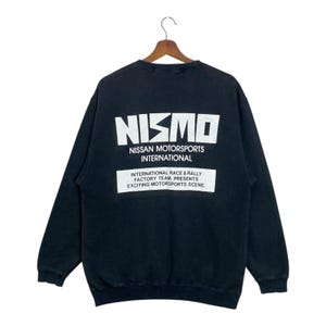 May include: Black sweatshirt with the white text "NISMO" and "NISSAN MOTORSPORTS INTERNATIONAL". Below the text is a white rectangle with the text "INTERNATIONAL RACE & RALLY FACTORY TEAM. PRESENTS EXCITING MOTORSPORTS SCENE."