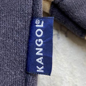 May include: Close-up of a navy blue fabric with a KANGOL label. The label is a dark blue rectangle with the white text "KANGOL" printed vertically. The fabric appears to be a sweatshirt material.