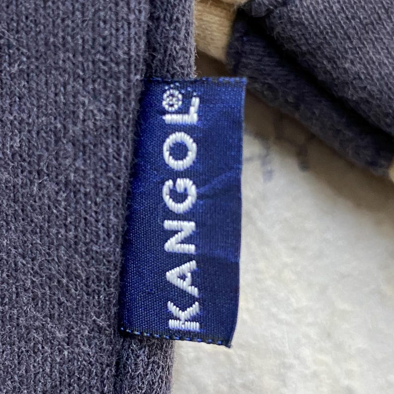 May include: Close-up of a navy blue fabric with a KANGOL label. The label is a dark blue rectangle with the white text "KANGOL" printed vertically. The fabric appears to be a sweatshirt material.