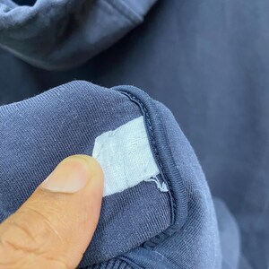 May include: Close-up of a navy blue hooded sweatshirt. A white label is attached to the inside collar. The fabric is a soft, knit material.