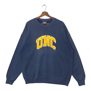 Op de afbeelding: Marineblauw sweatshirt met het woord "UNC" in gele blokletters. Het sweatshirt hangt aan een houten hanger. Het sweatshirt is een vintage stijl.