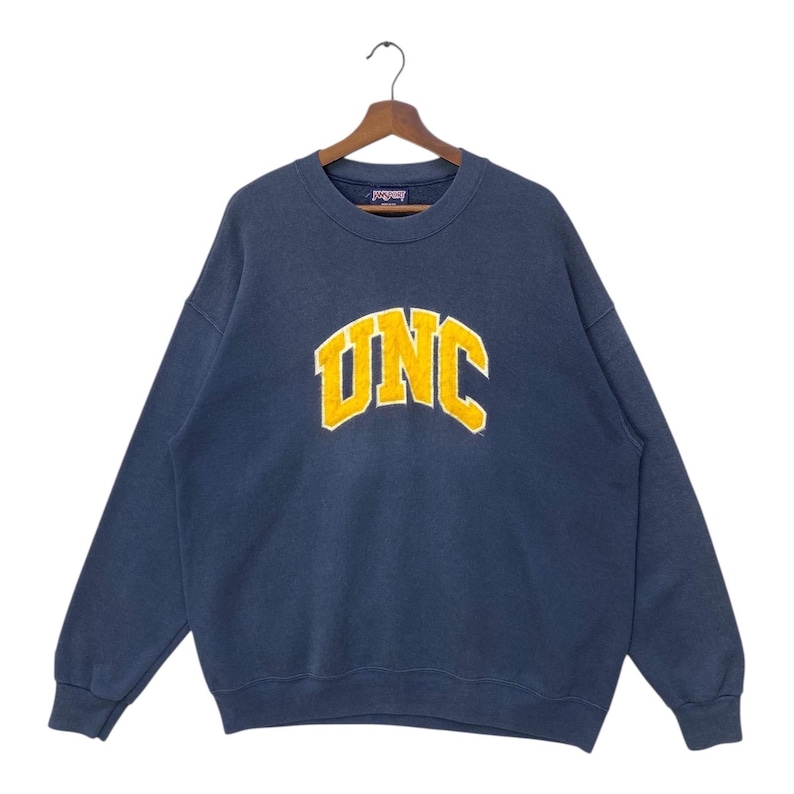 Op de afbeelding: Marineblauw sweatshirt met het woord "UNC" in gele blokletters. Het sweatshirt hangt aan een houten hanger. Het sweatshirt is een vintage stijl.