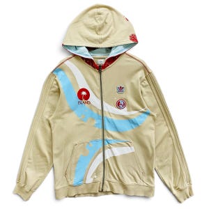 May include: A beige zip-up hoodie featuring a light blue and white curved graphic design on the front. The garment includes a red circular patch with a palm tree and the word ISLAND, an Adidas logo patch, and a red interior hood lining.