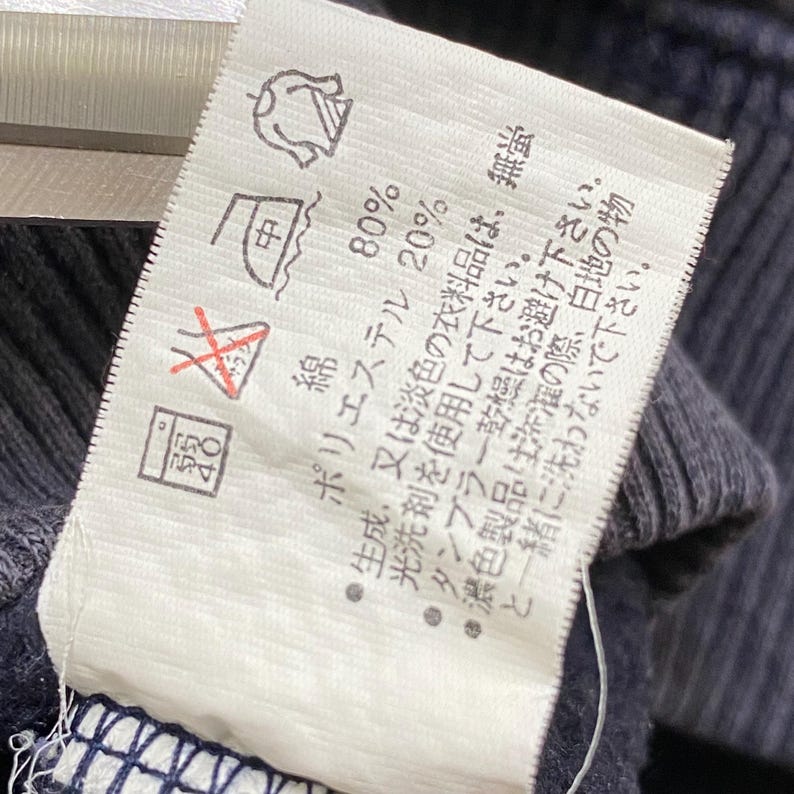 May include: A white clothing label with Japanese text and care symbols, attached to a dark blue fabric. The label details fabric composition and washing instructions, including percentages for cotton and polyester.