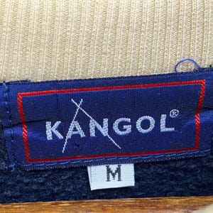 May include: A close-up shot of a navy blue label with the white text "KANGOL" and a stylized logo. The label has a red border and the letter "M" is visible below the brand name. The background is a tan fabric.