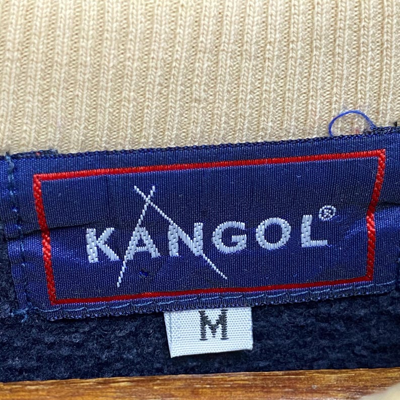 May include: A close-up shot of a navy blue label with the white text "KANGOL" and a stylized logo. The label has a red border and the letter "M" is visible below the brand name. The background is a tan fabric.