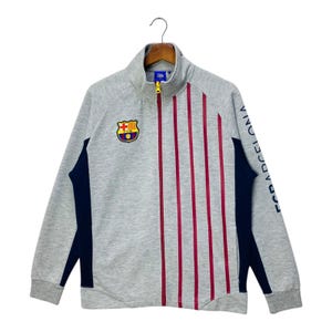 May include: A grey zip-up track jacket with a high collar. The jacket features a team logo on the chest, vertical red stripes, and navy blue accents. The text "FC BARCELONA" is printed on the sleeve.