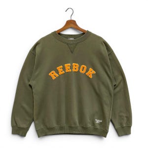 May include: An olive green Reebok sweatshirt with orange, arched lettering spelling out 'REEBOK'. The crew neck sweatshirt has long sleeves and a small Reebok logo on the lower right. It is displayed on a wooden hanger.
