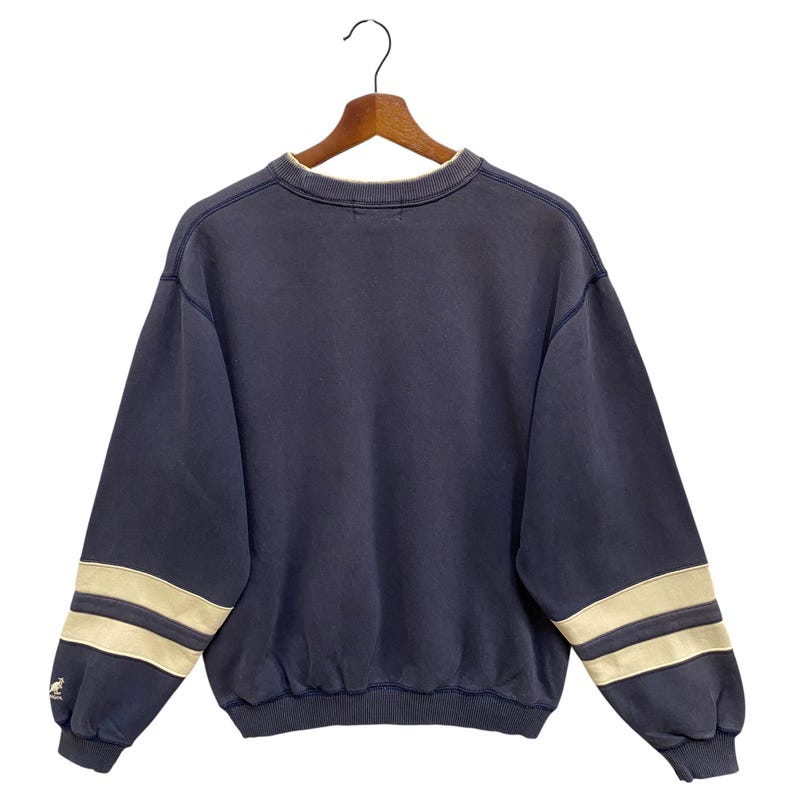 May include: Navy blue sweatshirt with cream stripes on the sleeves. The crew neck sweatshirt has a small logo on the left sleeve. The garment is suspended on a wooden hanger against a white backdrop.