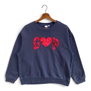 Könnte beinhalten: Ein marineblaues Langarm-Sweatshirt mit Rundhalsausschnitt und dem Schriftzug GAP, bei dem ein rotes Herz den Buchstaben A ersetzt. Die Buchstaben und das Herz haben eine strukturierte, schimmernde rote Oberfläche auf einem Holzkleiderbügel.