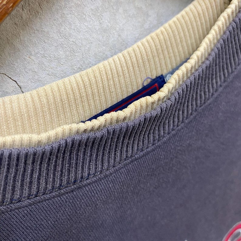 May include: Close-up of a vintage blue sweatshirt collar with a cream-coloured ribbed inner collar. A blue and red tag is visible between the collars. The fabric has a textured appearance.