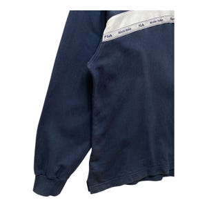 May include: Navy blue sweatshirt with a white and grey panel across the chest. The panel features the brand name "Fila" and the text "Biella Italia" in a repeating pattern. The sweatshirt has a ribbed cuff.