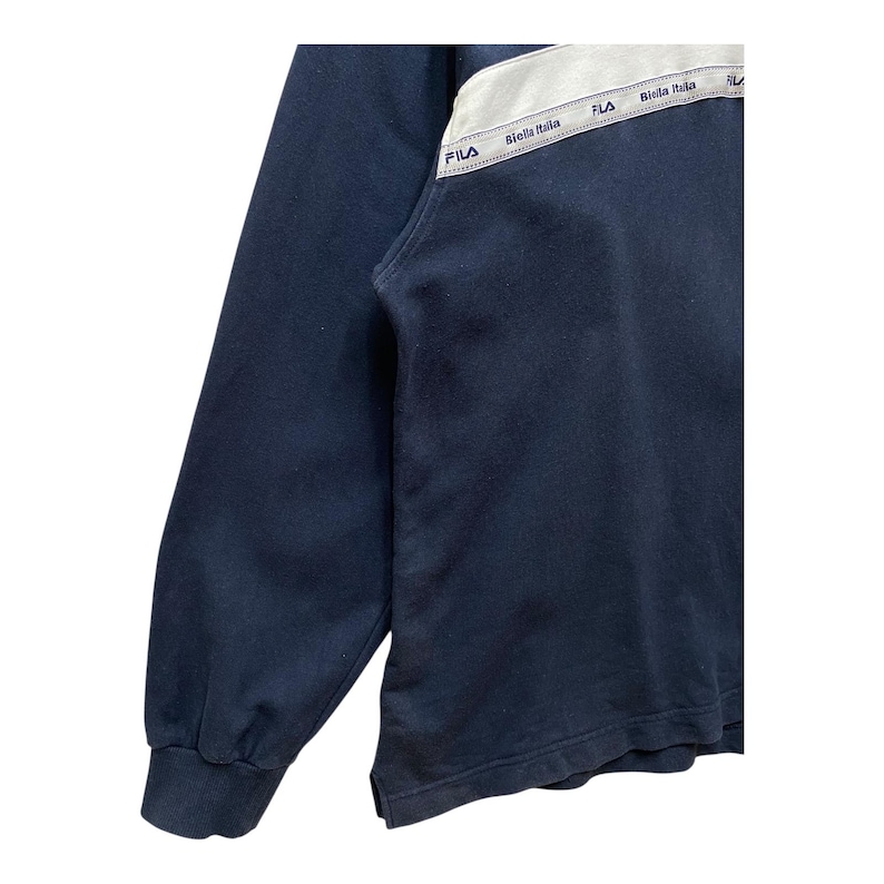 May include: Navy blue sweatshirt with a white and grey panel across the chest. The panel features the brand name "Fila" and the text "Biella Italia" in a repeating pattern. The sweatshirt has a ribbed cuff.
