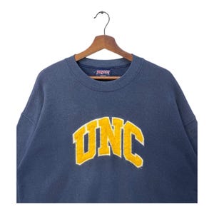 Op de afbeelding: Een marineblauw sweatshirt met het woord "UNC" in gele blokletters. Het sweatshirt hangt aan een houten kleerhanger. De merknaam "Jansport" is zichtbaar op de kraag.