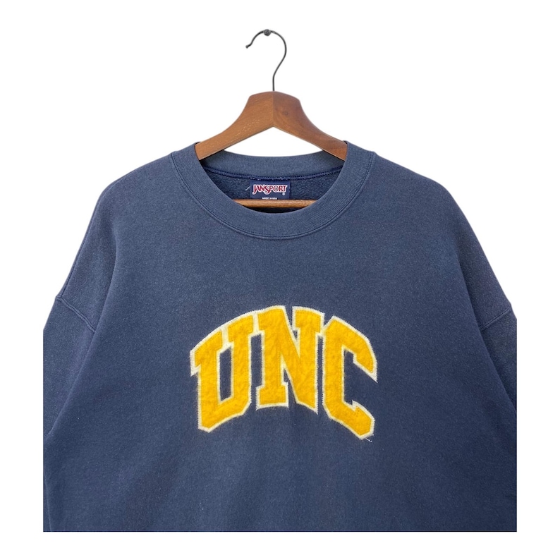 Op de afbeelding: Een marineblauw sweatshirt met het woord "UNC" in gele blokletters. Het sweatshirt hangt aan een houten kleerhanger. De merknaam "Jansport" is zichtbaar op de kraag.