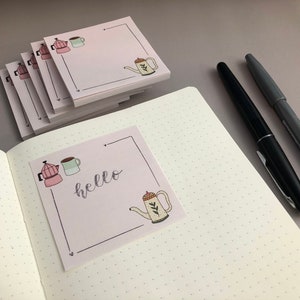 Puede incluir: Notas adhesivas rosas con ilustraciones de caf&eacute;: cafetera, taza y tetera. Una nota muestra la palabra "hello" en cursiva. Dos bol&iacute;grafos, uno negro y otro gris, descansan sobre un cuaderno punteado.