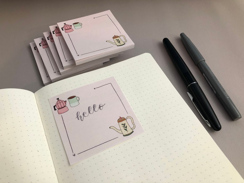 Puede incluir: Notas adhesivas rosas con ilustraciones de caf&eacute;: cafetera, taza y tetera. Una nota muestra la palabra "hello" en cursiva. Dos bol&iacute;grafos, uno negro y otro gris, descansan sobre un cuaderno punteado.
