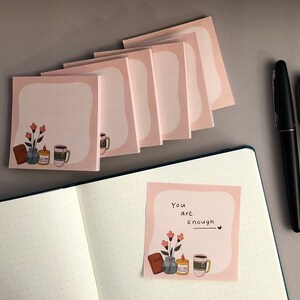 May include: Pink square sticky notes with a white border and illustrations of flowers, a mug, and a candle. One note has the text "You are enough." A notebook, black pen, and gray marker are also present.