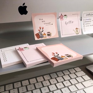 May include: Pink notepads with illustrations of coffee, flowers, and the words "You are enough" and "hello." One notepad has a list for "Saturday." The notepads are on a desk with a keyboard and computer.