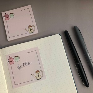 Puede incluir: Notas adhesivas rosas con ilustraciones con tem&aacute;tica de caf&eacute; y la palabra "hello". Un bol&iacute;grafo negro y un rotulador gris est&aacute;n junto a las notas y un cuaderno punteado abierto. Las ilustraciones incluyen una cafetera, una taza y una tetera.