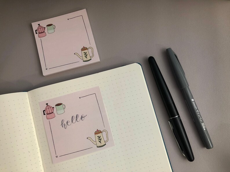 Puede incluir: Notas adhesivas rosas con ilustraciones con tem&aacute;tica de caf&eacute; y la palabra "hello". Un bol&iacute;grafo negro y un rotulador gris est&aacute;n junto a las notas y un cuaderno punteado abierto. Las ilustraciones incluyen una cafetera, una taza y una tetera.