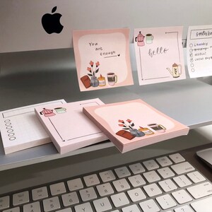 Puede incluir: Blocs de notas rosas con ilustraciones de cafeteras, tazas, flores y las palabras "You are enough" y "hello". Un bloc de notas tiene una lista para el "s&aacute;bado". Los blocs de notas est&aacute;n apilados en un escritorio cerca de un teclado.