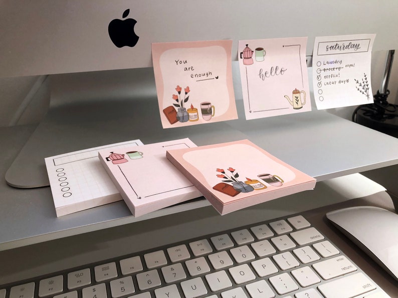 Puede incluir: Blocs de notas rosas con ilustraciones de cafeteras, tazas, flores y las palabras "You are enough" y "hello". Un bloc de notas tiene una lista para el "s&aacute;bado". Los blocs de notas est&aacute;n apilados en un escritorio cerca de un teclado.