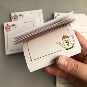 Puede incluir: Una pila de peque&ntilde;os blocs de notas cuadrados con una ilustraci&oacute;n dibujada a mano de una tetera. La tetera es de color crema con un asa y un pico marrones, y hojas verdes. Otros blocs de notas tienen ilustraciones de cafeteras y tazas.